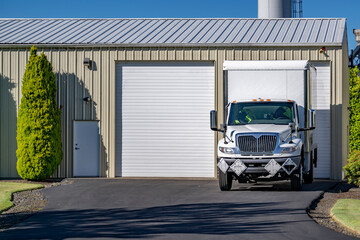 Local business day cab compact middle duty rig semi truck with box trailer standing on the industrial parking lot with workshop building
