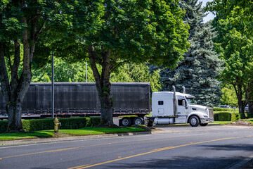 Powerful white big rig semi truck with extended cab transporting cargo in Conestoga dry van semi trailer turning on the local road from warehouse parking lot with green trees