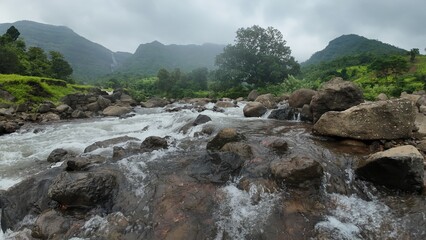 Stream flowing over small rocks with Shayadri hill in background, lush greenery during monsoon.