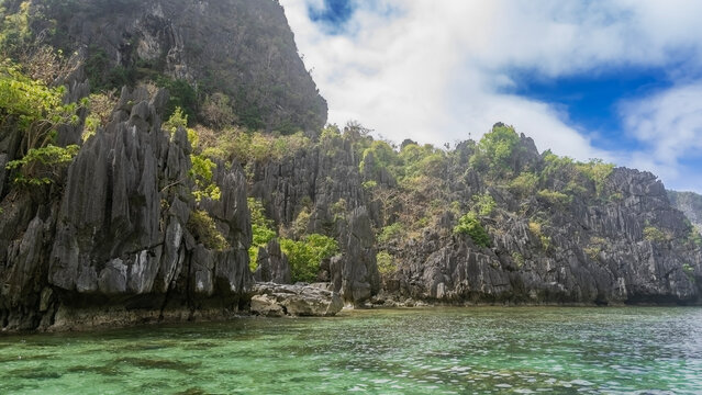 Picturesque karst rocks with steep slopes and sharp peaks surround the turquoise lagoon. Green tropical vegetation on the cliffs. Clouds in the blue sky. Philippines. Palawan.