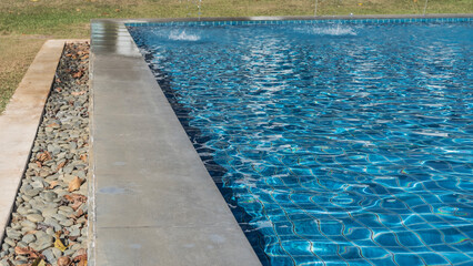 A fragment of a rectangular swimming pool. The blue tiled bottom is visible through the clear water. Ripples, undulations, highlights on the surface. Concrete barrier, pebbles around the perimeter. 
