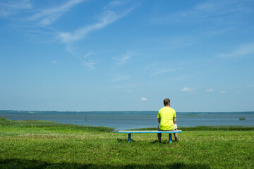 man sitting on a bench and looking at the water and grass in front of him, with blue sky and clouds, stock photo, depth of field