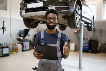 Confident auto mechanic smiling with clipboard and thumbs up in repair workshop. Professional...