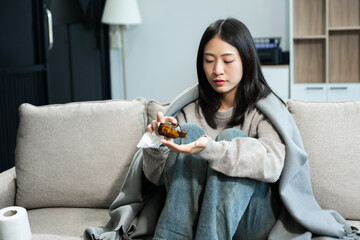 Naklejka premium Young Asian woman suffering from flu symptoms, covered with a blanket on a sofa. Concept of illness, healthcare, and recovery