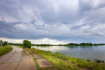A paved path winds through lush green grass and leads toward a calm river with a distant forest line. The sky is filled with dramatic, swirling clouds, suggesting an approaching storm..