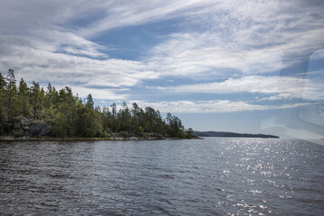 A tranquil view across a still lake with a shoreline of dense trees. The sky is bright and cloud-filled, reflecting on the waters surface.