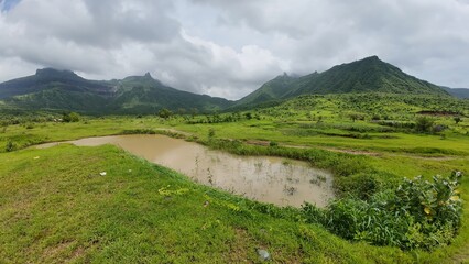 Obraz premium Scenic View of Sahyadri Hills with Pond, Lush Green Meadow, and Cloudy Sky during Monsoon Season
