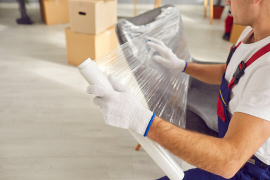 Young male mover packing furniture preparing for moving in the living room. Man worker from professional moving service wrapping chair with plastic wrap. Move and relocation concept.