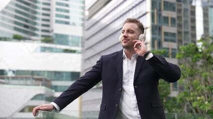 Professional businessman wearing headphone while listening music at rooftop. Caucasian project manager moving to relaxing music while looking at skyscraper and stand at green city. Lifestyle. Urbane. - Powered by Adobe