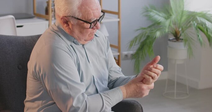 Retired old senior man, elderly person alone at home, suffering from arthritis and holding, rubbing his painful hand. Feeling bad problems with joints, fingers, wrist pain, rheumatoid inflammation