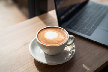 Close-up of Hot coffee latte with latte art milk foam in cup mug and laptop computer on wood desk office desk in coffee shop at the cafe,during business work concept