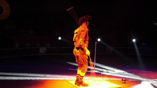 A clown with a funny hairstyle entertains guests in the circus arena in the light of colorful spotlights and smoke. He's tap dancing. Circus performance.