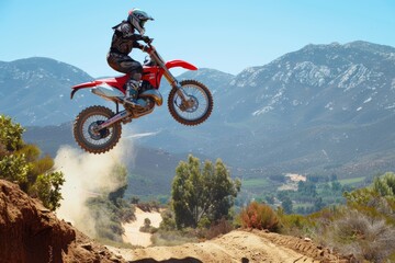 Naklejka premium A dirt bike rider performing a high jump over a rugged, dusty trail with mountains and a clear blue sky in the background