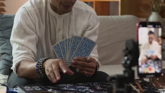 Cheerful man tarot reader doing predicting online and recording video with smartphone in living room