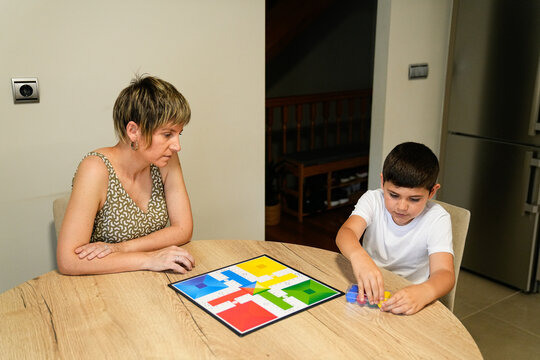 Mother watching her son play Parcheesi at the dining table, deeply engaged in the game.