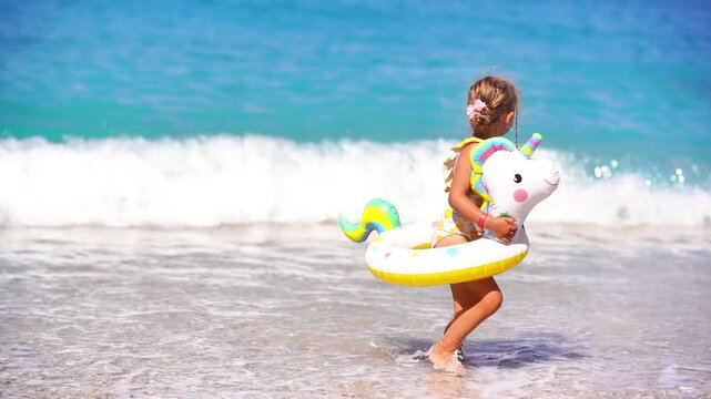 Summer vacation scene of a 5-year-old blonde girl walking on the beach with a swim ring around her waist, turquoise sea in the background. Capturing the joy and freedom of childhood during a sunny bea