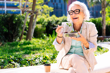 middle age entrepreneur woman laughing happy while eating a takeaway salad sitting outdoors in a city park, concept of healthy food and active lifestyle in adulthood, copyspace for text