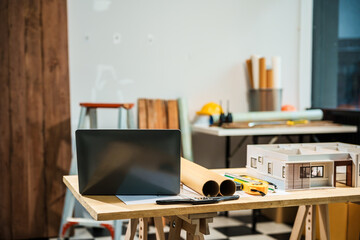 A rear view of a laptop on a desk, displaying an architect's model of a house with various rooms, house plans, and wooden textures for a renovation project.