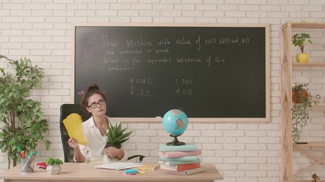 Charismatic woman college teacher with funny hairstyle sitting at desk in classroom holding pot with plant and waving fan. Education concept.