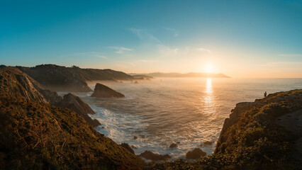 Fototapeta premium The Loiba cliffs at sunset with the sun casting a golden light. A person is seen in the distance admiring the view. The sky is filled with soft, wispy clouds.