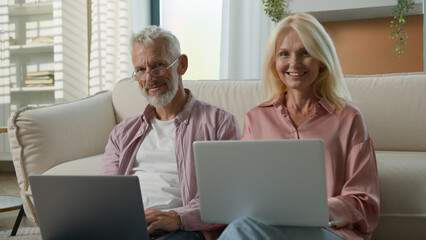 Happy Caucasian elderly married couple senior man woman retired businesspeople business work on Internet together in living room using laptops smiling looking camera older people and modern technology