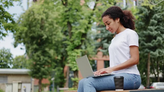 Tired young african american woman gently tapping keys of personal laptop while appreciating fresh outdoors air. Attentive exhausted female worker sitting on wooden bench and focused on tasks.