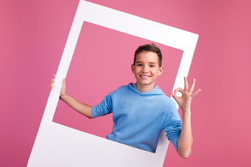 Photo portrait of cute teen boy hold instant photo frame okey approve dressed stylish blue clothes isolated on pink color background