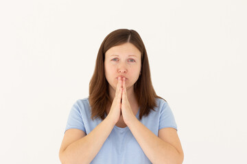 Portrait of imploring girl tilts head, purses lips, looks sadly at camera, makes praying gesture, asks for help and support, begs boyfriend for apologize, wears blue t-shirt, isolated on white wall