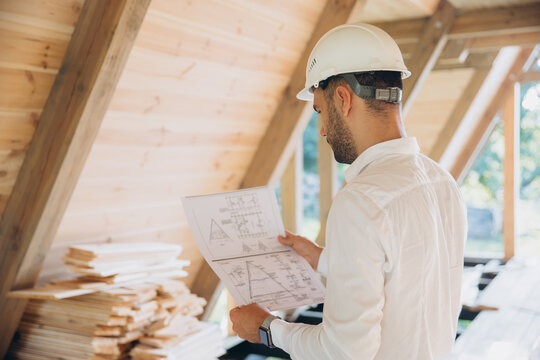 Architect wooden buildings. Man stands under frame wooden house. Man architect studies documentation or blueprints. Guy in protective helmet at construction site. Architect under timber framed house