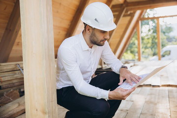 Architect wooden buildings. Man stands under frame wooden house. Man architect studies documentation or blueprints. Guy in protective helmet at construction site. Architect under timber framed house