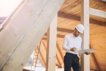 Fototapeta premium Architect wooden buildings. Man stands under frame wooden house. Man architect studies documentation or blueprints. Guy in protective helmet at construction site. Architect under timber framed house