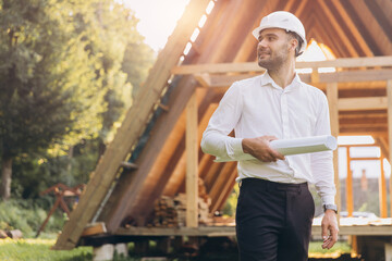 Construction engineer in white helmet. Developer with construction documentation on the background of wooden modular building under construction. Portrait of smiling architect near eco friendly house.