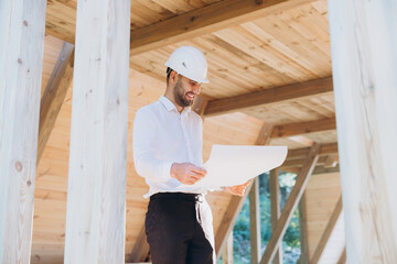 Construction engineer in white helmet. Developer with construction documentation on the background of wooden modular building under construction. Portrait of smiling architect near eco friendly house.