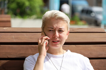 Girl with short blonde hair talking on mobile phone while sitting on street wooden bench. Tomboy...