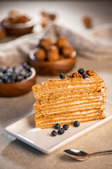 A slice of layered honey cake garnished with blueberries on a white plate, with more cake and a wooden bowl in the background
