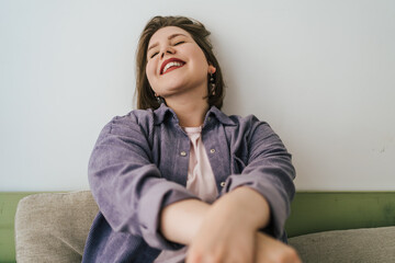 Cheerful plus-size woman sitting on couch, laughing with closed eyes. Relaxed, joyful expression, showcasing self-confidence and happiness. Body positivity and authentic emotions.