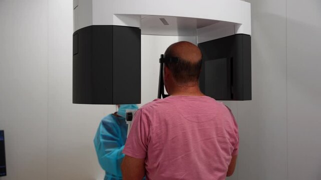 Female dentist in blue robe adjusts male patient's head and operates orthopantomography device for a panoramic radiography