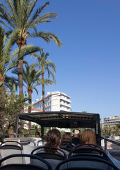 An open top tourist bus in Cannes in the south of France with a white building, blue sky and palm trees, from a tourist's point of view