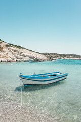 In Donousa, a Greek island in the Cyclades, a small boat peacefully anchors in clear, shallow waters surrounded by a brilliant blue sky and a coastline of white sand and rocky cliffs