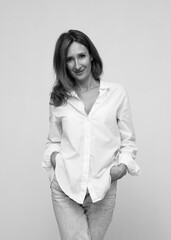 Young attractive woman with wavy red hair and freckles wearing white shirt and jeans, with hands in pocket,  smiling and looking at camera. Black and white image.