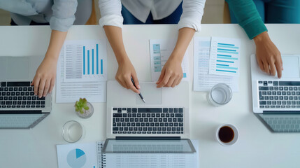 A top down view of a team collaborating on business reports and charts at a desk, using laptops and analyzing data together in a modern office setting.