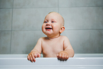 Baby boy bathes at home in a white bathtub with water. Baby smiling and happy splashing in the water at home.