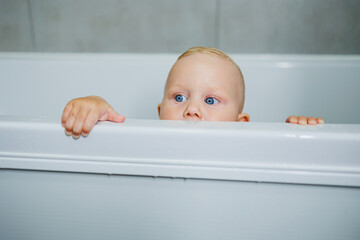 Baby boy bathes at home in a white bathtub with water. Baby smiling and happy splashing in the water at home.