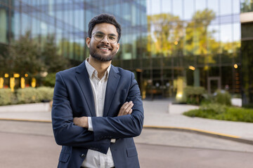 Confident young businessman standing outside office building with arms crossed and smiling. Professional wearing glasses and suit jacket, representing success, confidence, and leadership