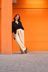 Woman Leaning Against Orange Wall in Casual Outfit. smiling at the camera.