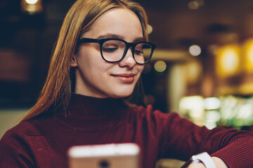 Positive woman checking time on wearable electronic wristwatch while waiting friend in cafe and communicated via mobile phone, successful hipster girl in eyeglasses looking at smartwatch screen