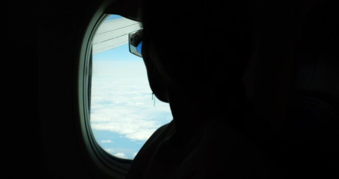 Woman dark silhouette near rounded window with light clouds view outside in commercial airplane. Female passenger with glasses travels by plane. Flight abroad