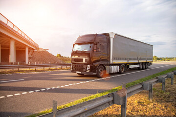 a black modern truck with a semi-trailer is driving along the road against the backdrop of a bridge and sunset. Freight transportation services, business. Copy space for text