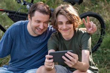 Father and teenager son taking funny selfie photo together on mobile phone during summer rest in park outdoor