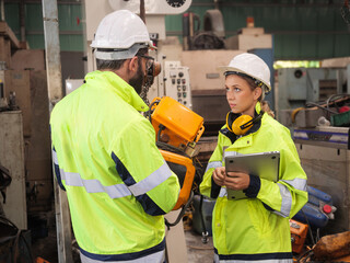 Confident young female mechanical engineer in a protective uniform, helmet while listening to colleague explanation workflow the CNC Machinery while working at the plant.